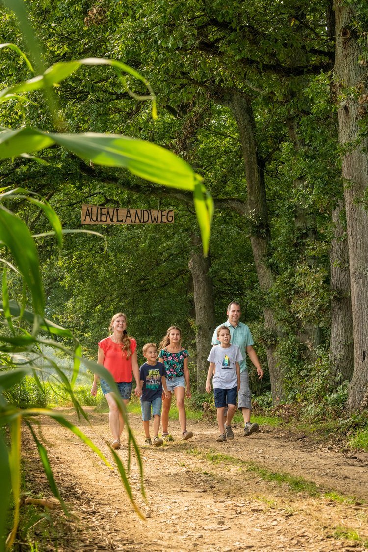 Eine Familie läuft einen Waldweg entlang, über dem das Schild "Auenlandweg" hängt