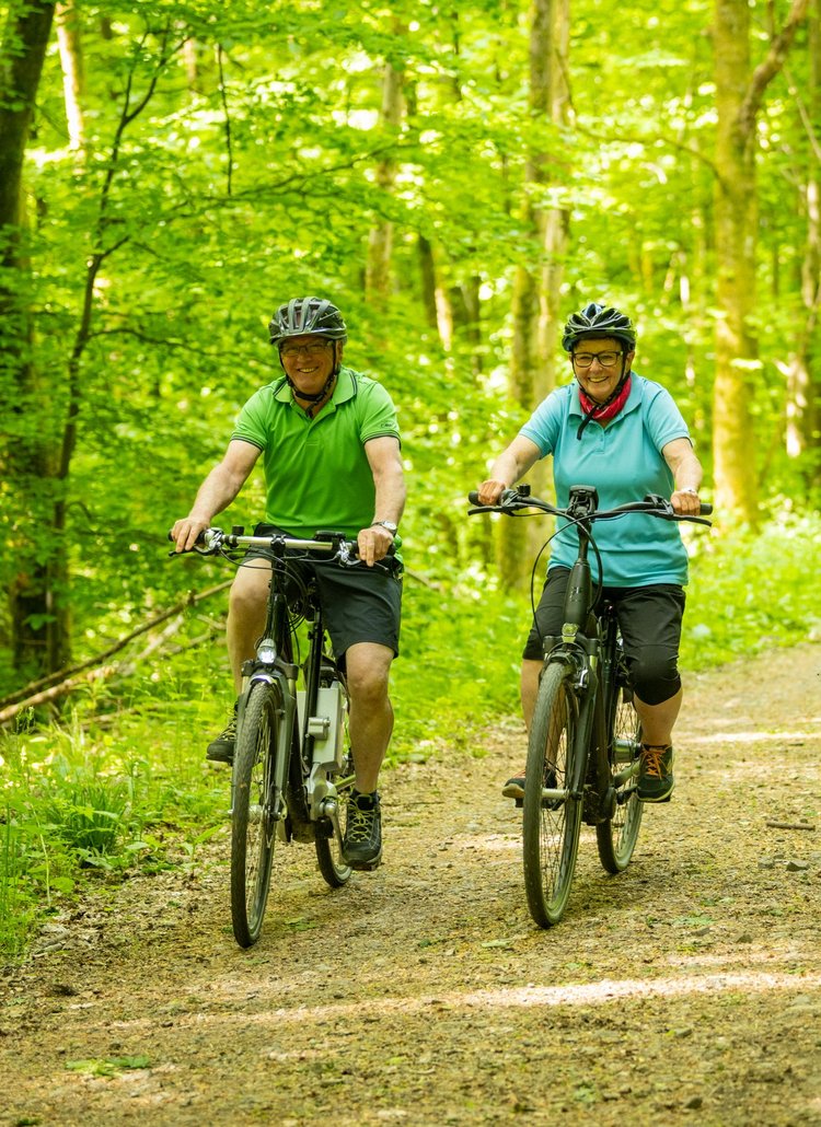 3 ältere Personen sind lachend mit dem Fahrrad auf einem Waldweg unterwegs.