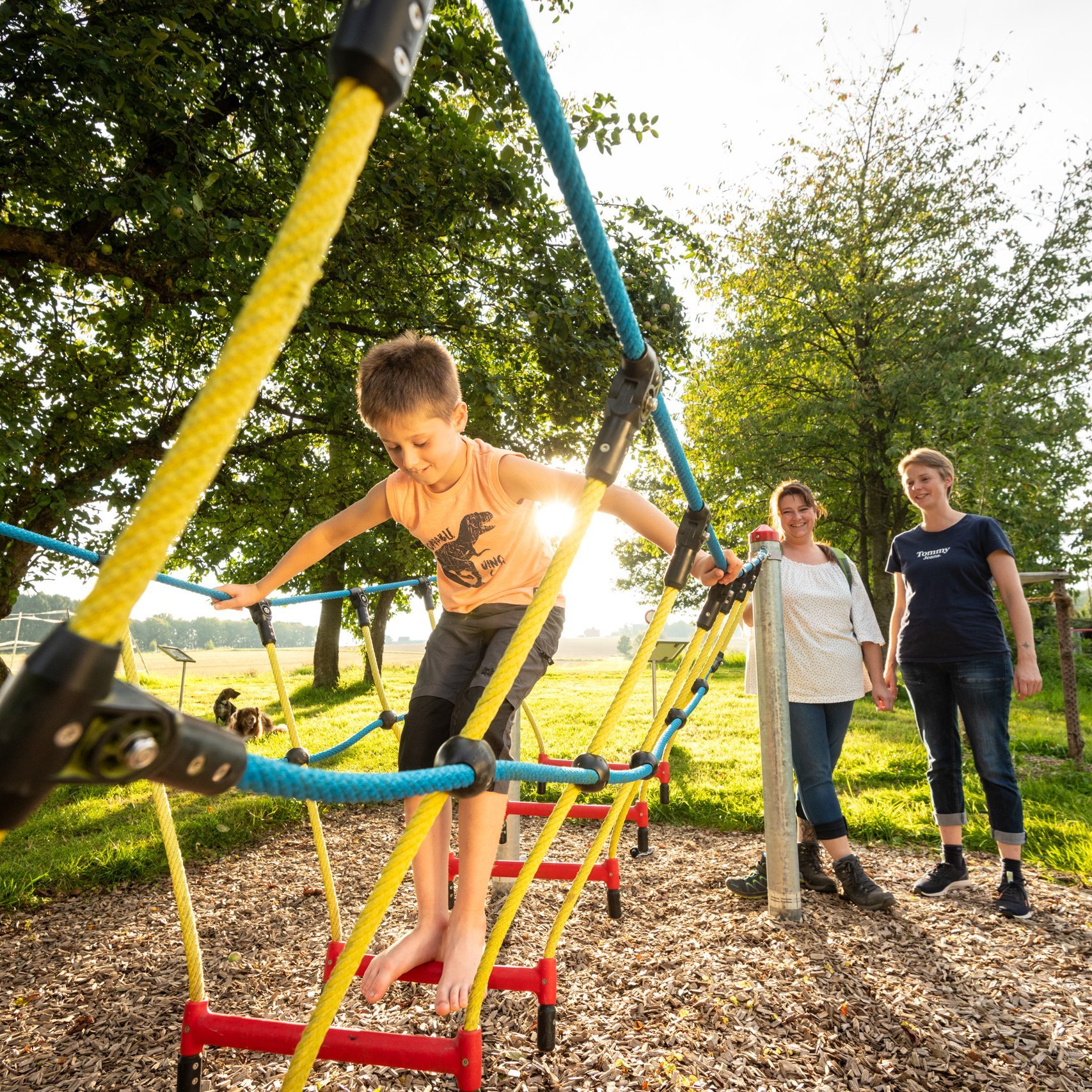 Ein Junge klettert auf einem Seilgerüst auf einem Spielplatz und wird von zwei Frauen beobachtet
