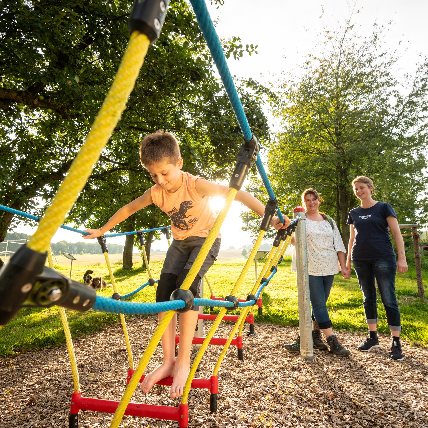 Ein Junge klettert auf einem Seilgerüst auf einem Spielplatz und wird von zwei Frauen beobachtet