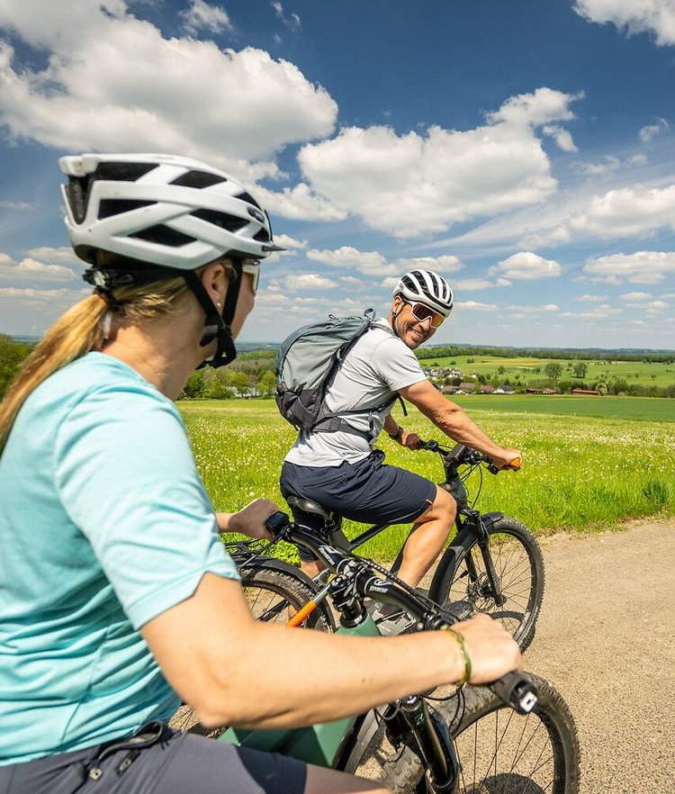 Ein Mann auf dem Fahrrad schaut zurück auf eine Frau hinter ihm. Im Hintergrund sieht man weite Wiesen und Wälder des Westerwaldes.