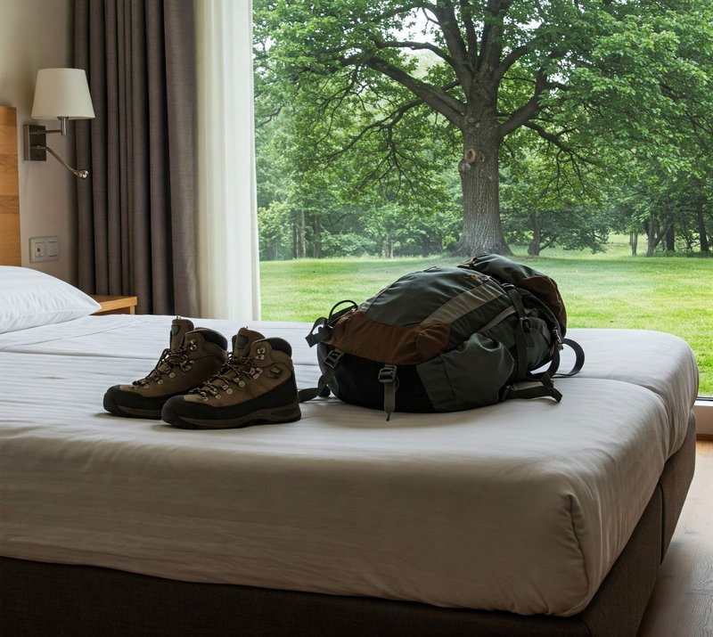 Rucksack und Wanderschuhe auf einem Hotelbett vor einem großen Fenster, dass in die Natur blickt.