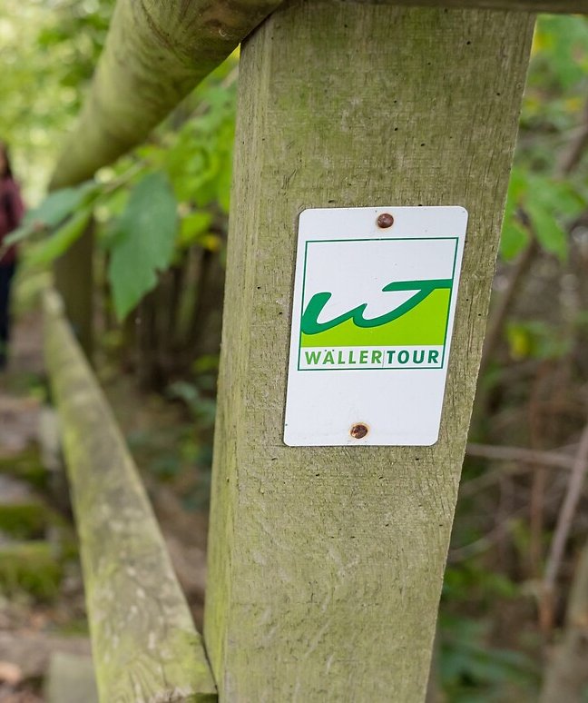 An einem Treppengeländer ist die Markierung der Wäller Touren angebracht, das ein grünes W in einem gerahmten weißen Untergrund zeigt. 