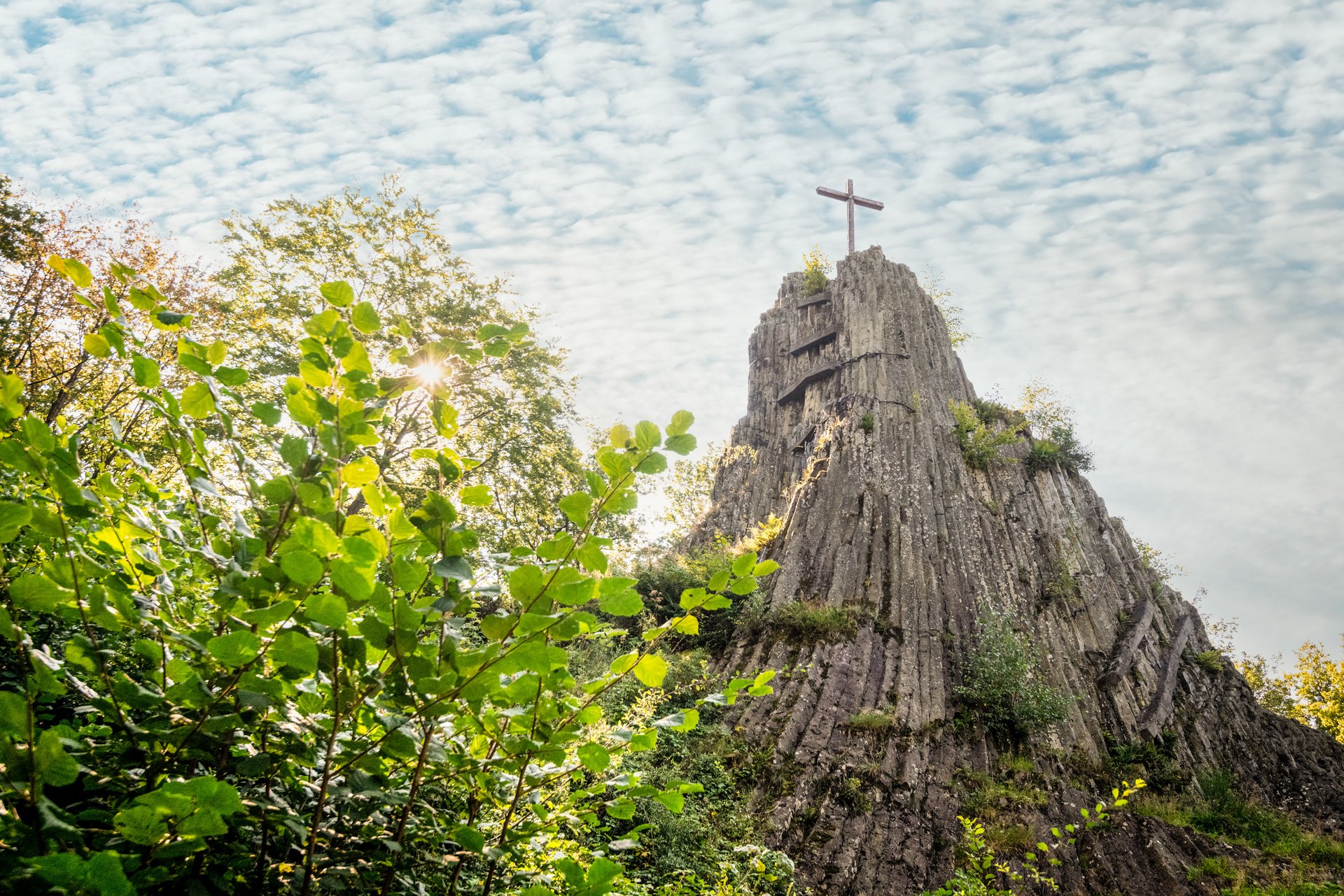 Zwischen Bäumen ragt das Gipfelkreuz des Druidenstein auf den Basaltformationen vor wolkigen Himmel empor. 