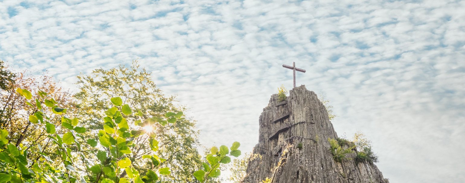 Zwischen Bäumen ragt das Gipfelkreuz des Druidenstein auf den Basaltformationen vor wolkigen Himmel empor. 