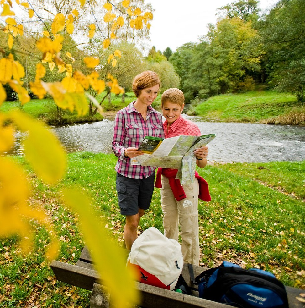 Zwei Wanderer rasten im Herbst an einer Bank am Fluss und schauen in die Wanderkarte