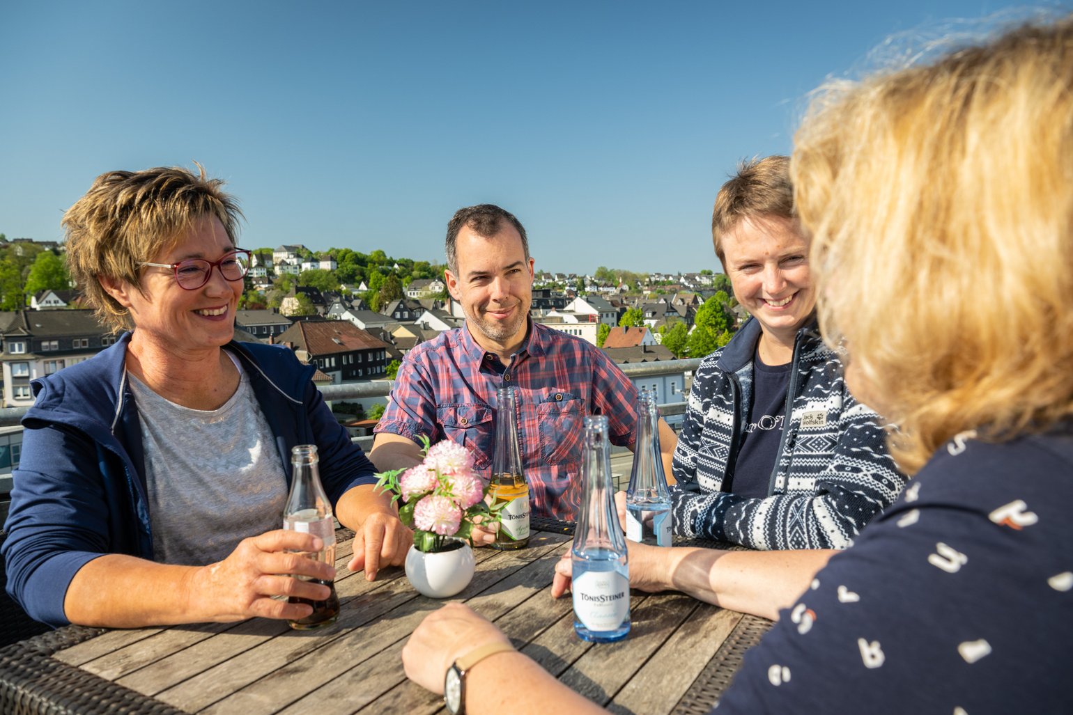 Eine Gruppe Freunde lacht bei einer Pause im Sonnenschein mit Getränken. Im Hintergrund sieht man den Ausblick über Wissen.