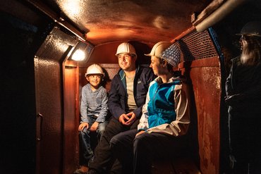 Ein Vater sitzt mit zwei Kindern mit Helmen in der Grubenbahn des Schaubergwerks bei schummerigen Licht 