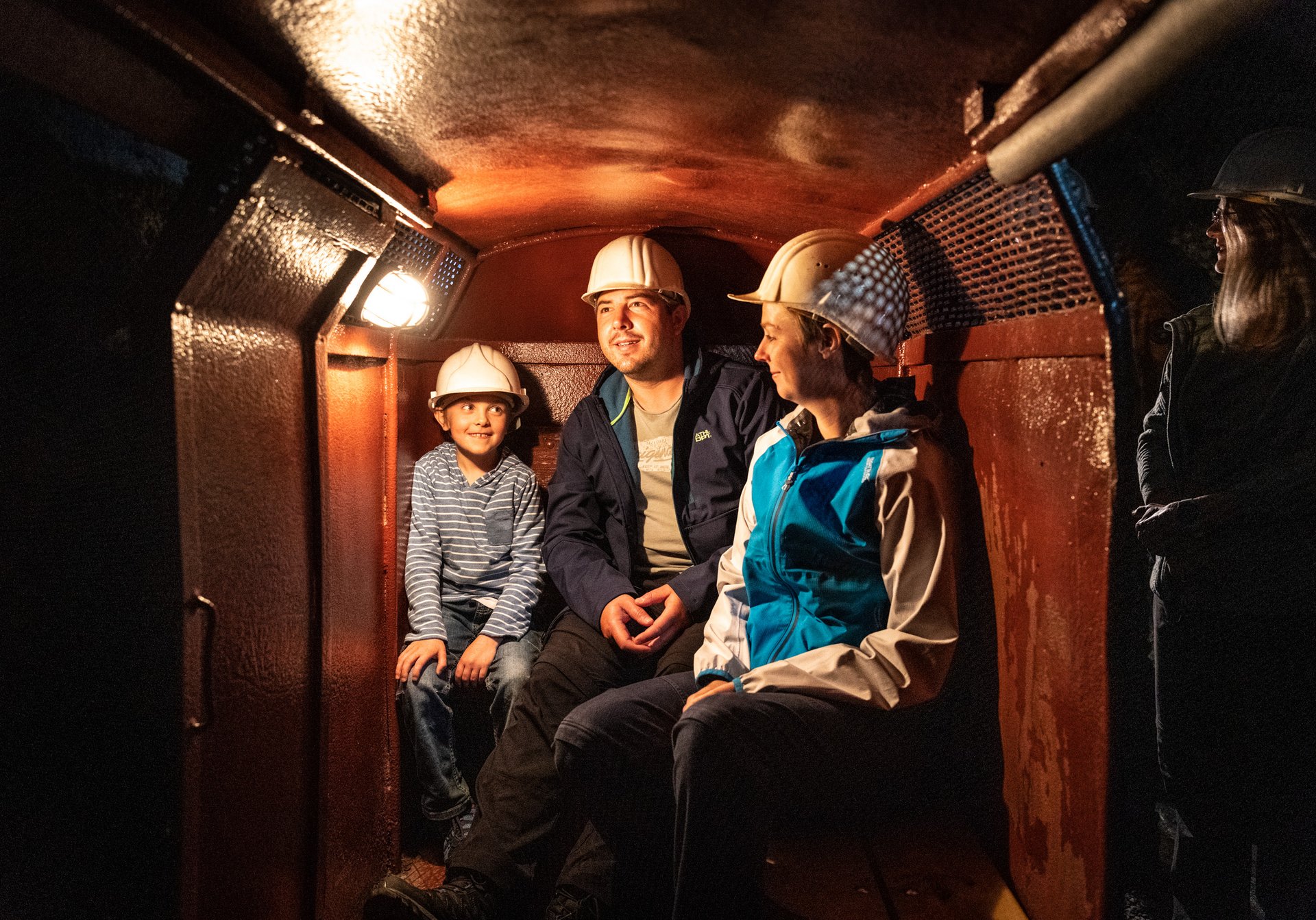 Ein Vater sitzt mit zwei Kindern mit Helmen in der Grubenbahn des Schaubergwerks bei schummerigen Licht 