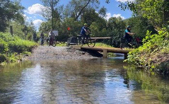 Eine Reihe Radfahrer überqueren einen Bach über einen Holzsteg