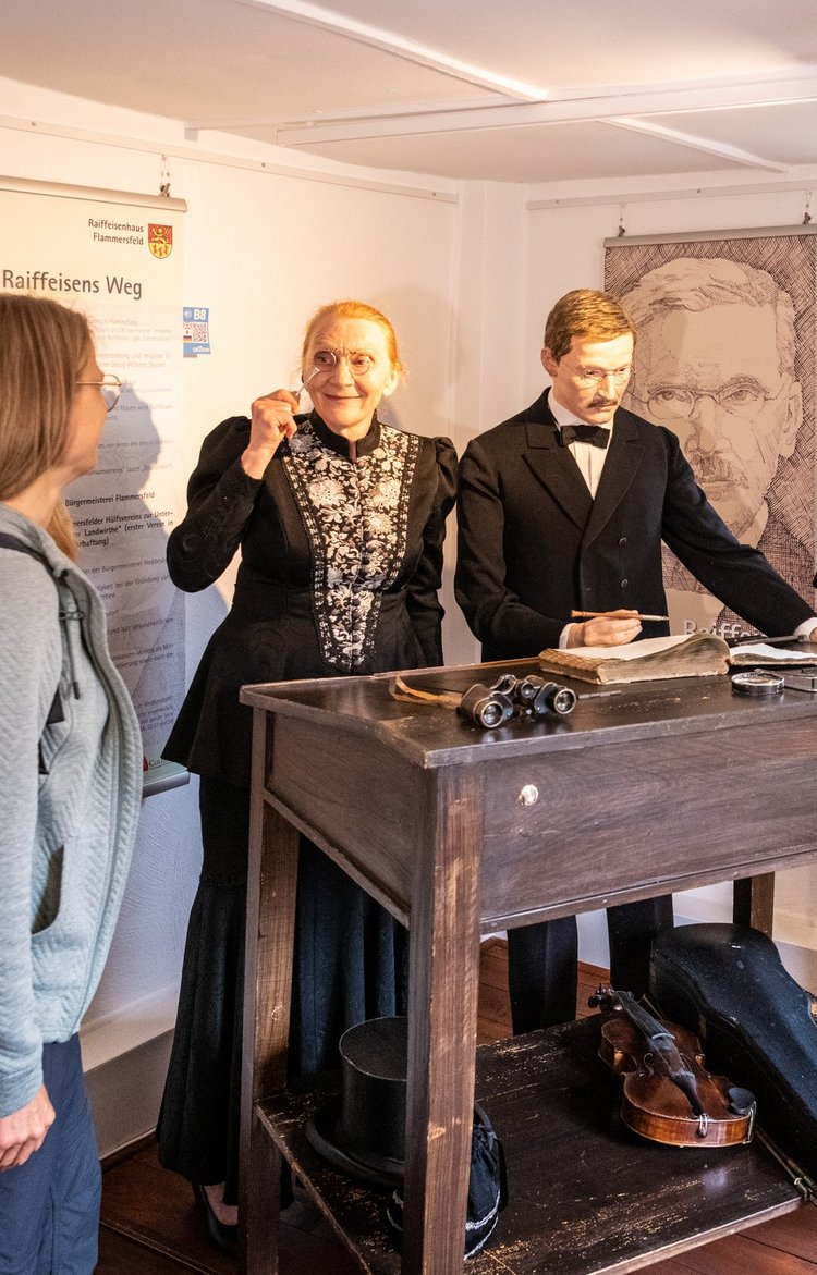 Im Raiffeisenhaus Hamm besucht eine Gruppe die Wachsfigur von Friedrich Wilhelm Raiffeisen und die Gästeführerin gewandet in den Kleidern aus der Zeit.