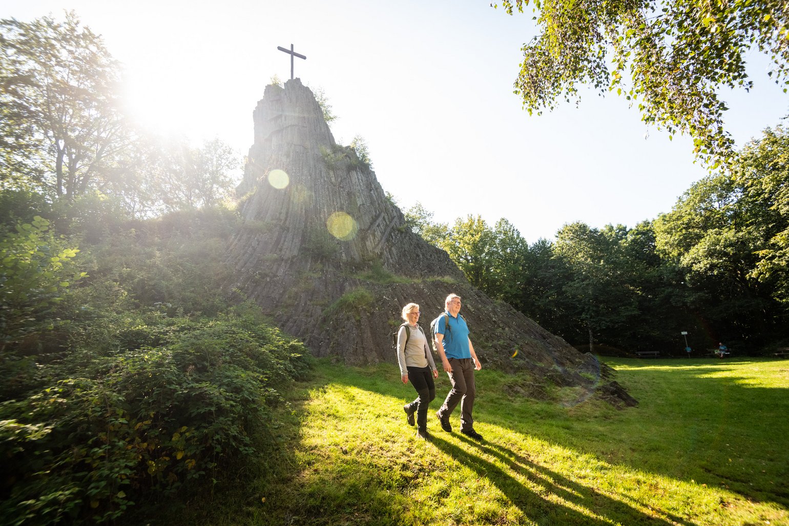 Zwei Wanderer laufen entlang des Druidensteins, der sich im Hintergrund auftürmt, Richtung Wald. Die Sonne geht hinter dem Druidenstein unter. 