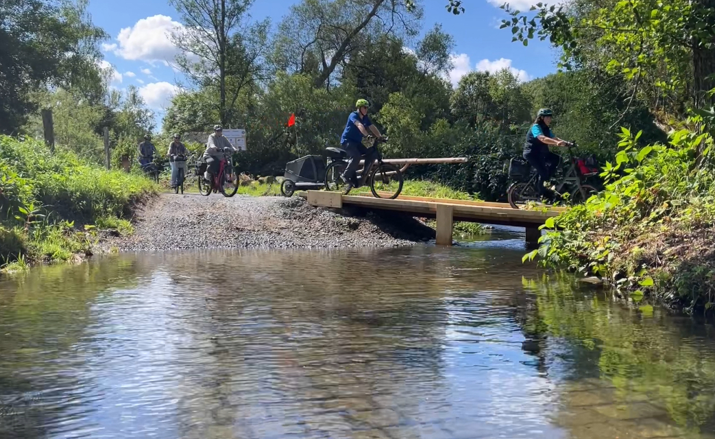 Eine Reihe Radfahrer überqueren einen Bach über einen Holzsteg