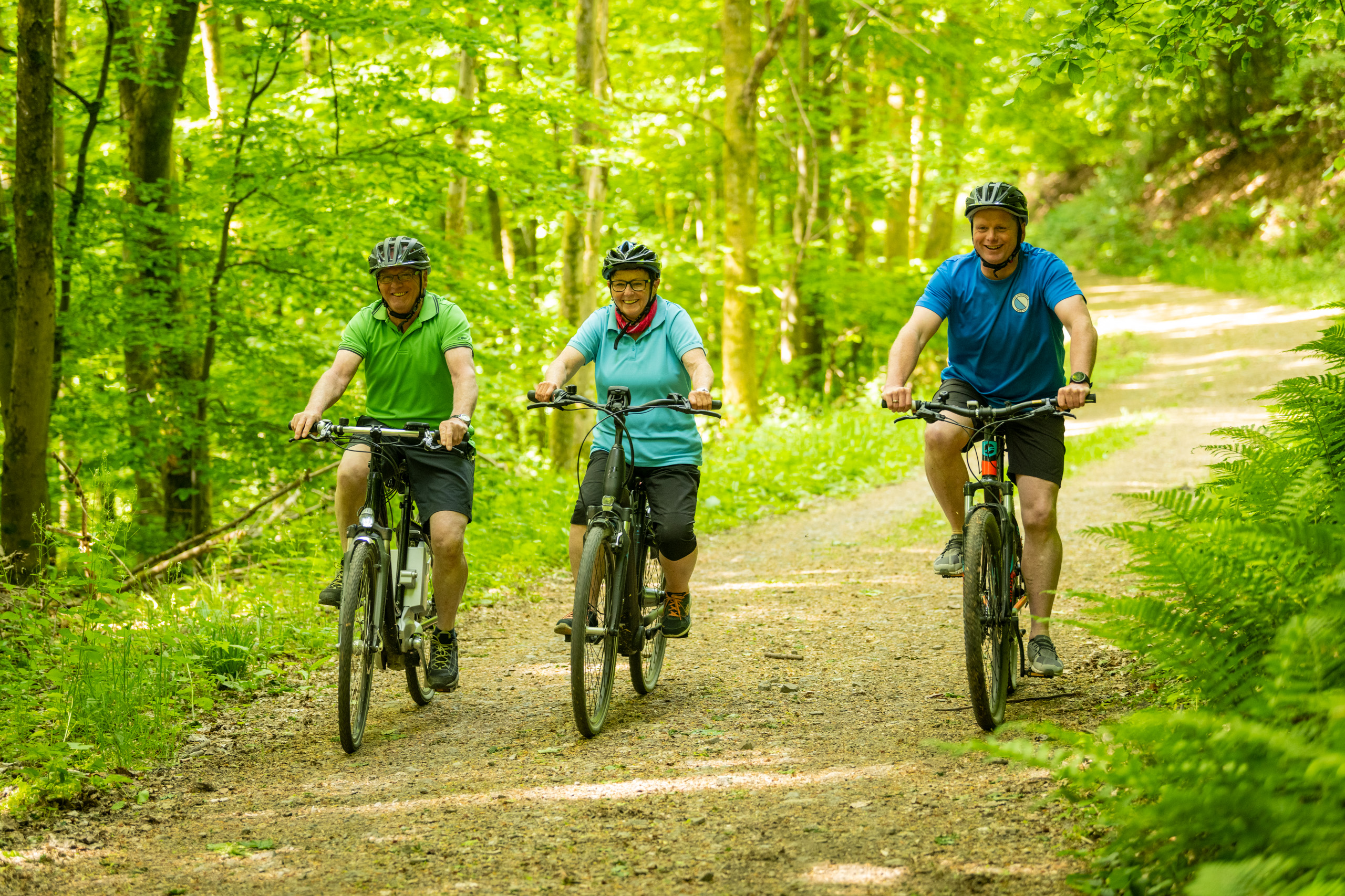 3 ältere Personen sind lachend mit dem Fahrrad auf einem Waldweg unterwegs.