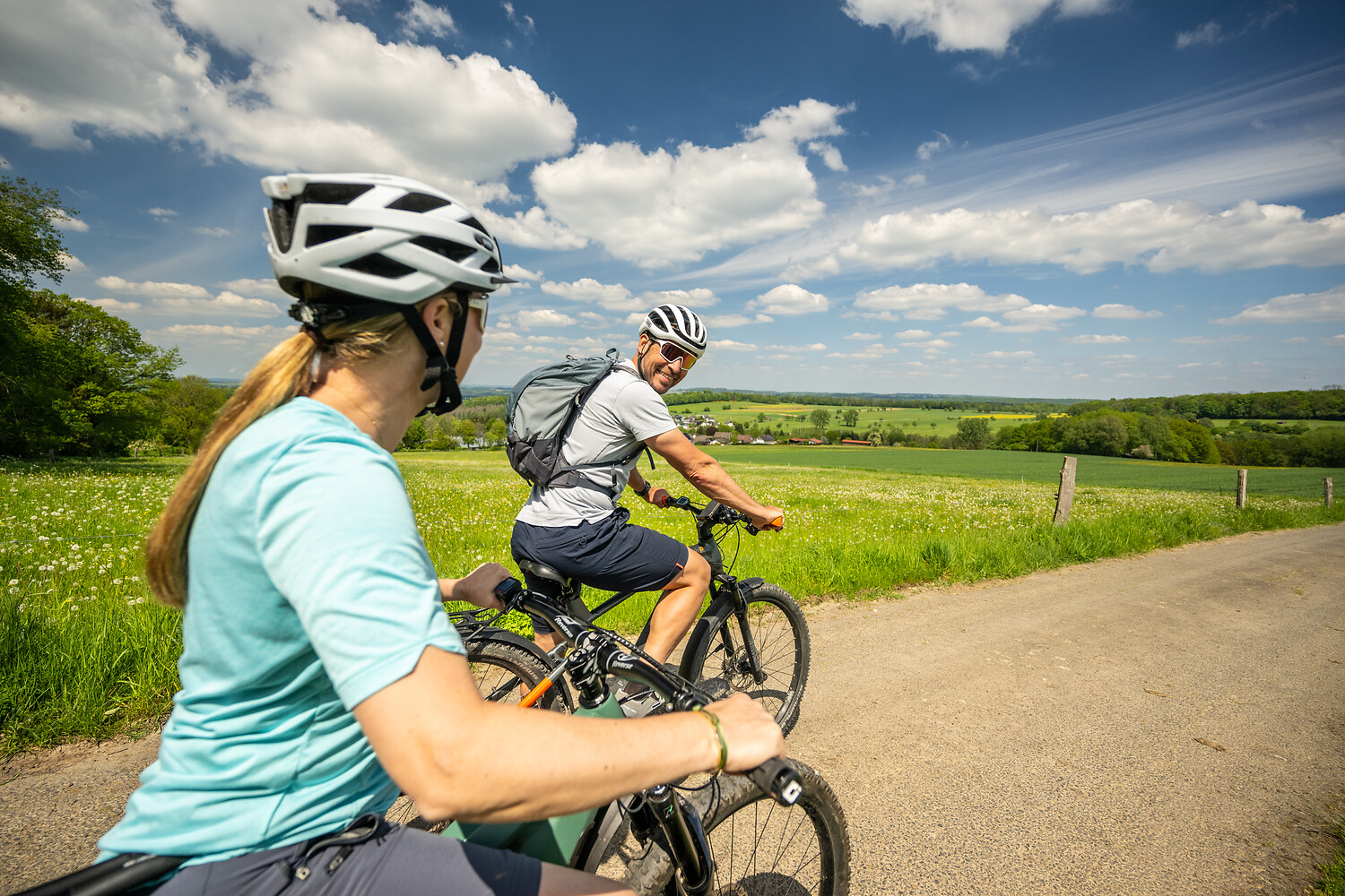 Ein Mann auf dem Fahrrad schaut zurück auf eine Frau hinter ihm. Im Hintergrund sieht man weite Wiesen und Wälder des Westerwaldes.