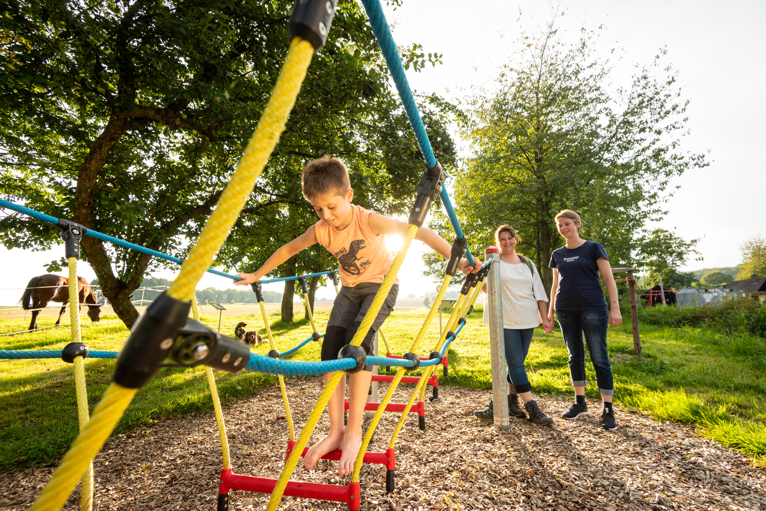 Ein Junge klettert auf einem Seilgerüst auf einem Spielplatz und wird von zwei Frauen beobachtet